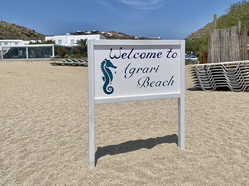 A sign for Agrari Beach in Paralia Agrari, Mykonos, Greece, with a sandy beach in the background.