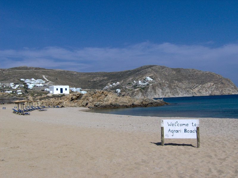 A serene Agrari Beach in April with calm waters and scattered rocks along the shore.