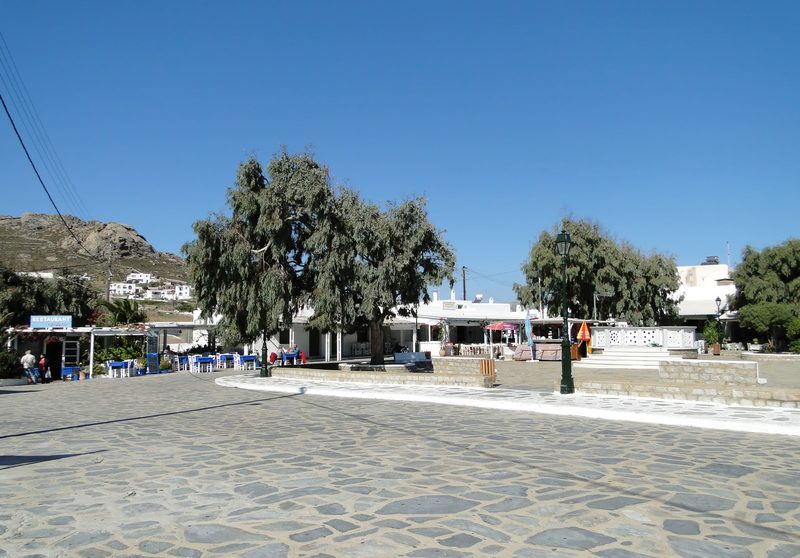 A bustling square in Ano Mera Village, Mykonos, with traditional architecture and people enjoying the sunny day.