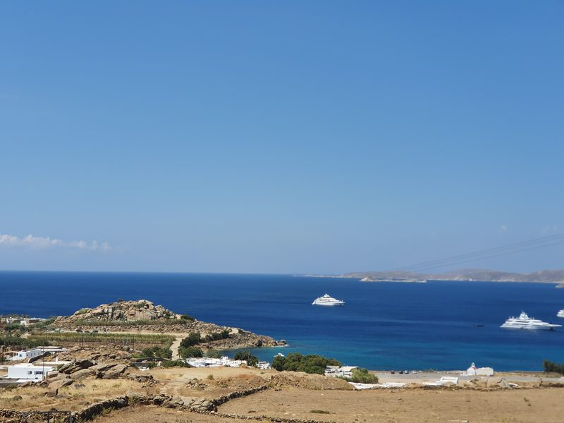 A south coast bay near Mykonos seen from a headland, with yachts anchored in the blue water below