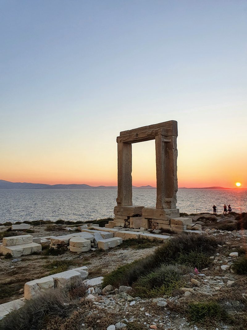 The Portara silhouetted against an orange sunset sky, with the sun setting into the sea behind it