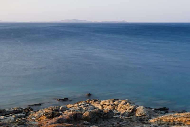 A long-exposure shot of waves crashing on a rocky shore with Mykonos in the distance, Tinos, Greece.
