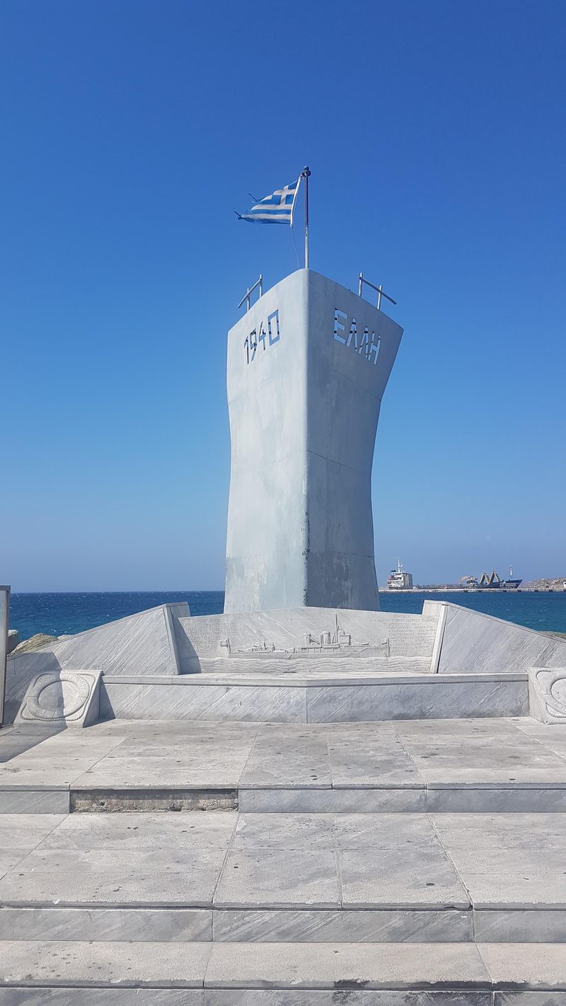 The marble war memorial on the waterfront at Tinos port, flying the Greek flag