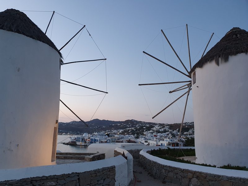 Looking between the wooden sail frames of two Mykonos windmills toward the town and sea at dusk