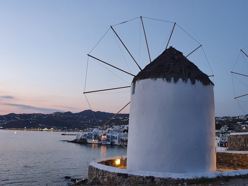 A Kato Mili windmill illuminated at dusk with the lights of Little Venice and Mykonos harbour behind it