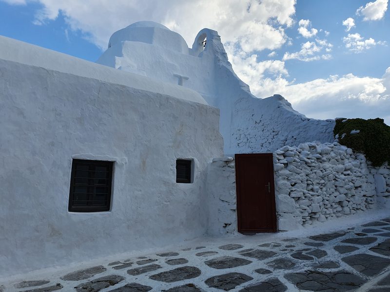 A side view of Panagia Paraportiani's stacked whitewashed chapels above a stone wall and cobbled lane