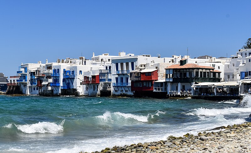 Colorful waterfront buildings of Little Venice in Mykonos with waves lapping against their foundations under a clear blue sky